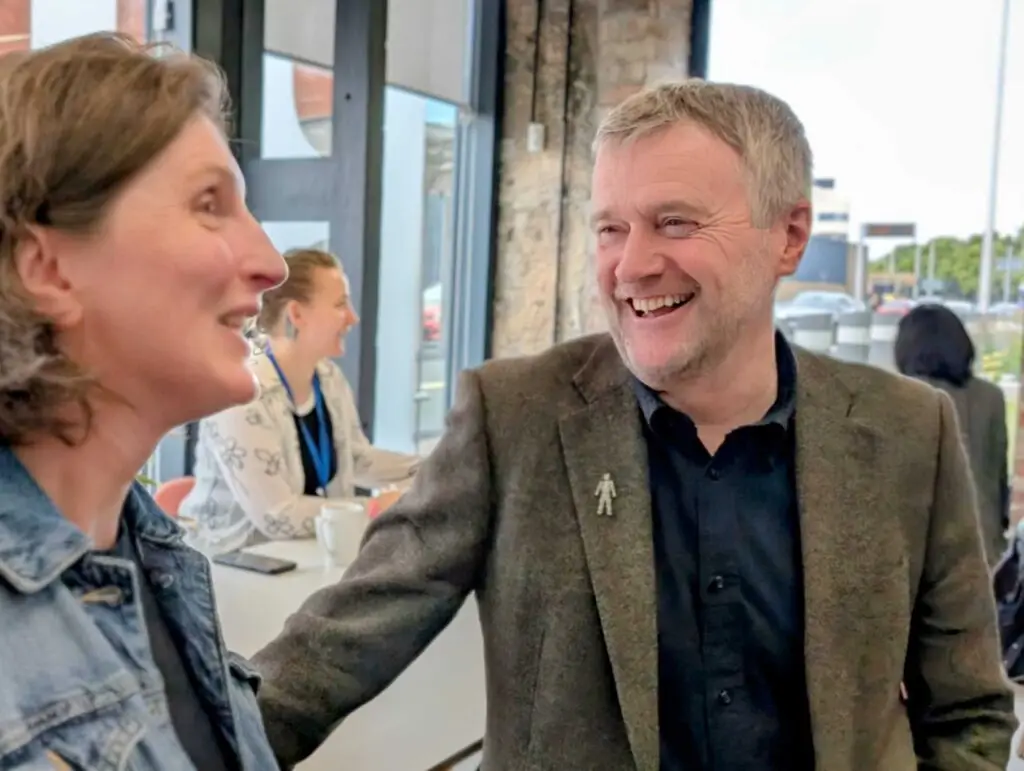 David smiles talking to Hilary in a conference setting