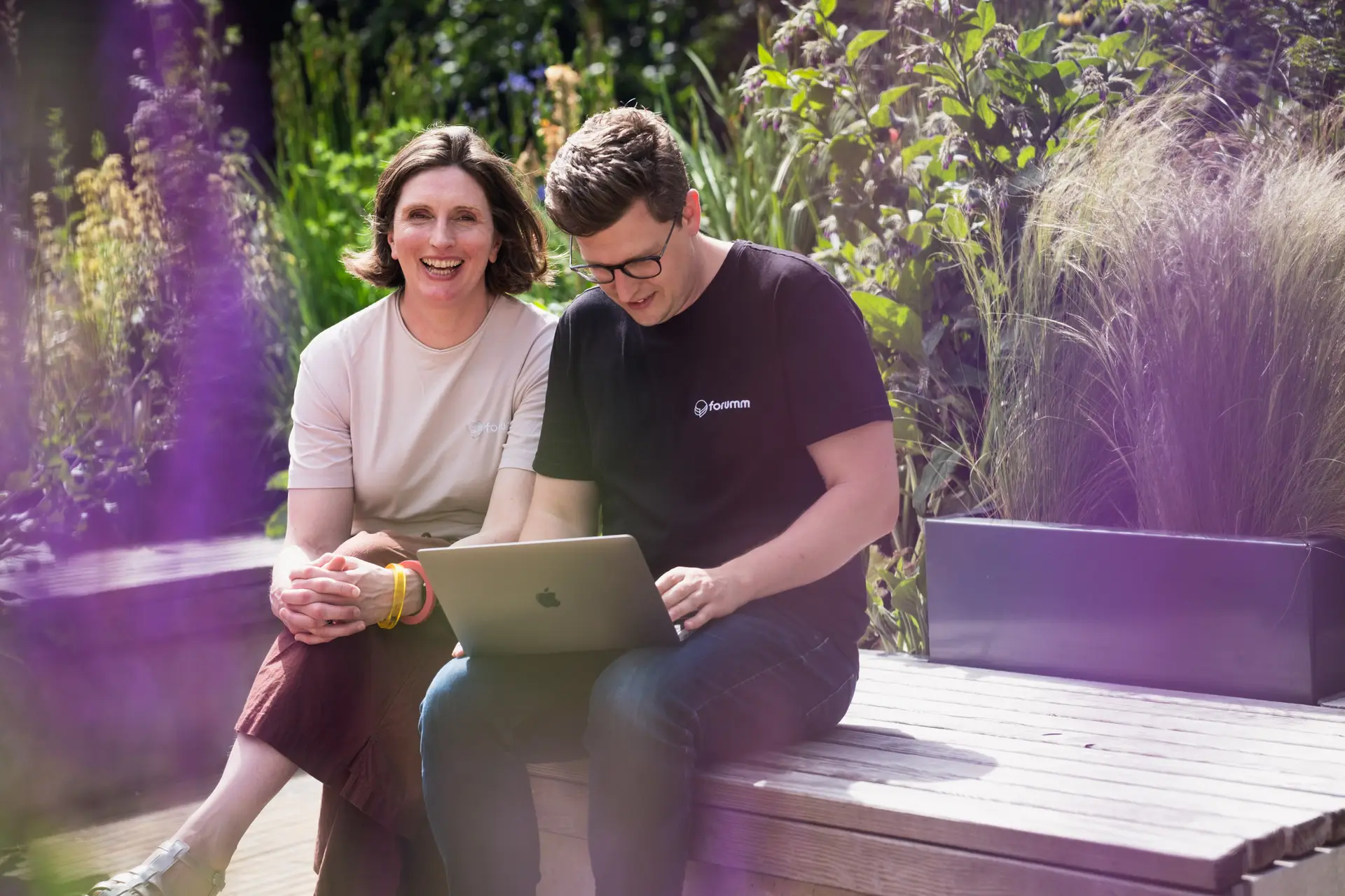 Woman smiles at camera while man works on computer in a garden
